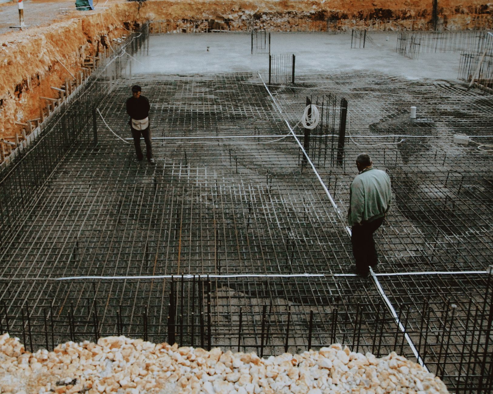 Two workers inspecting a rebar grid foundation at a construction site.