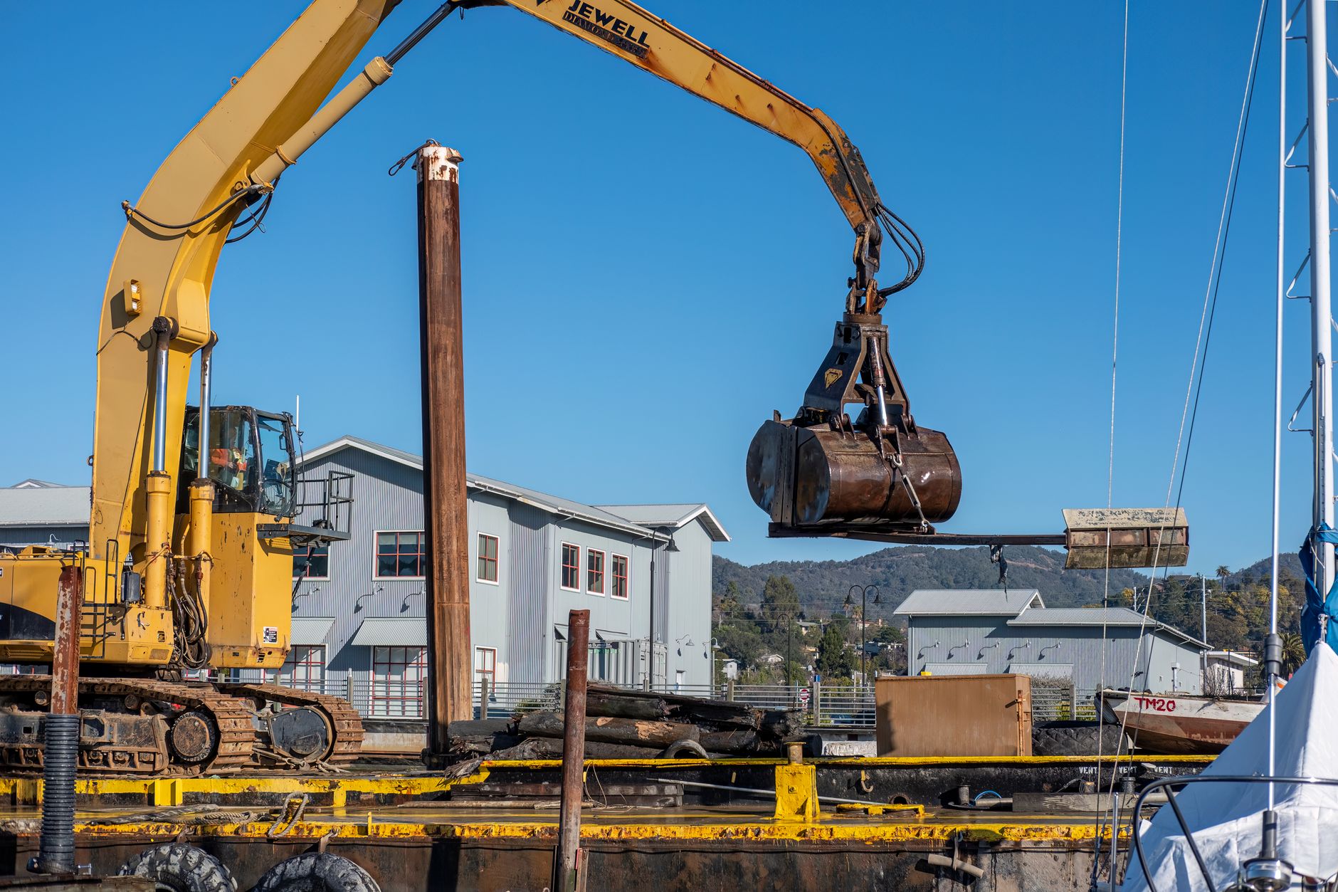 Yellow excavator with crane bucket at a construction site near buildings.