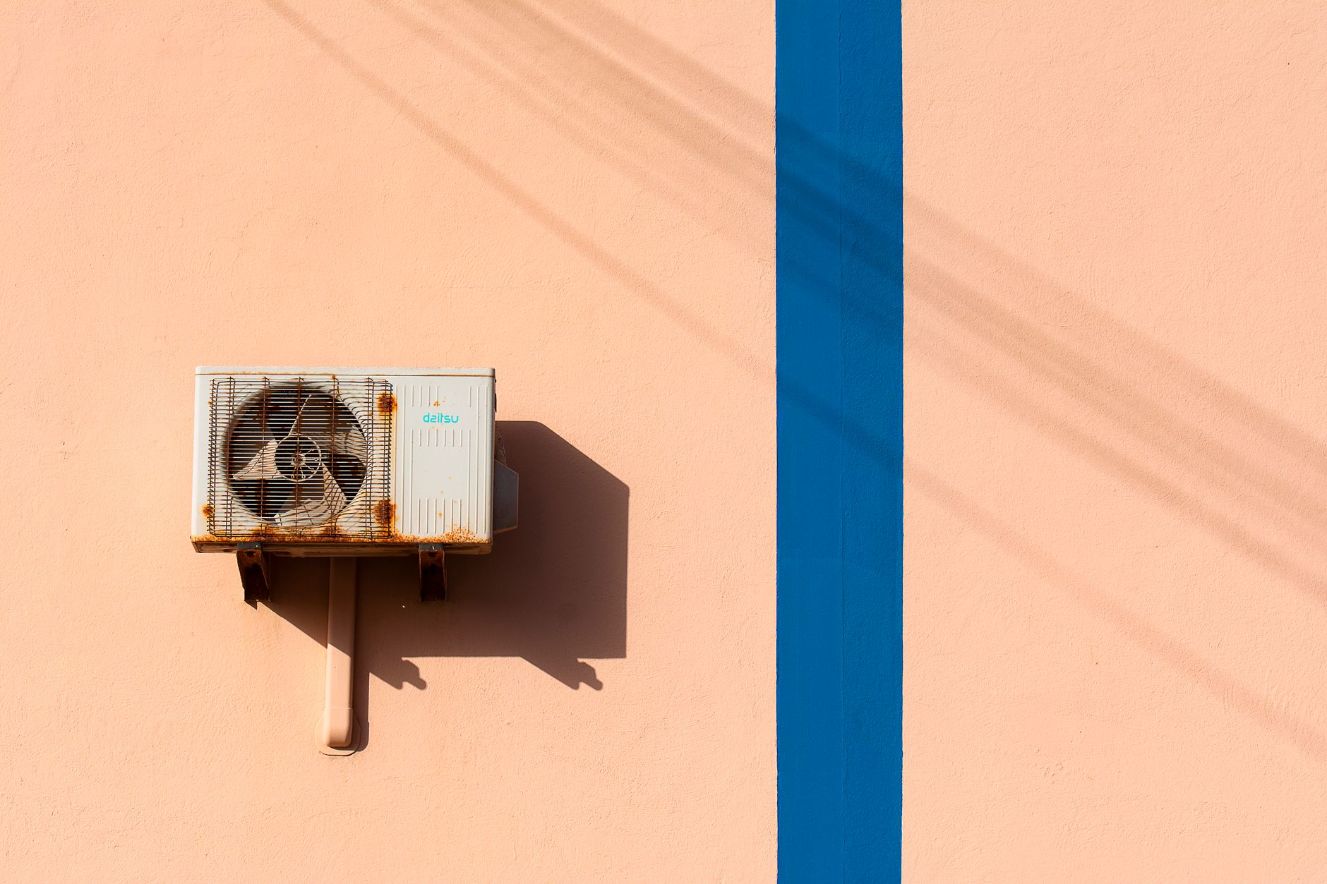 Rusty air conditioning unit casting shadow on a peach wall with blue stripe.