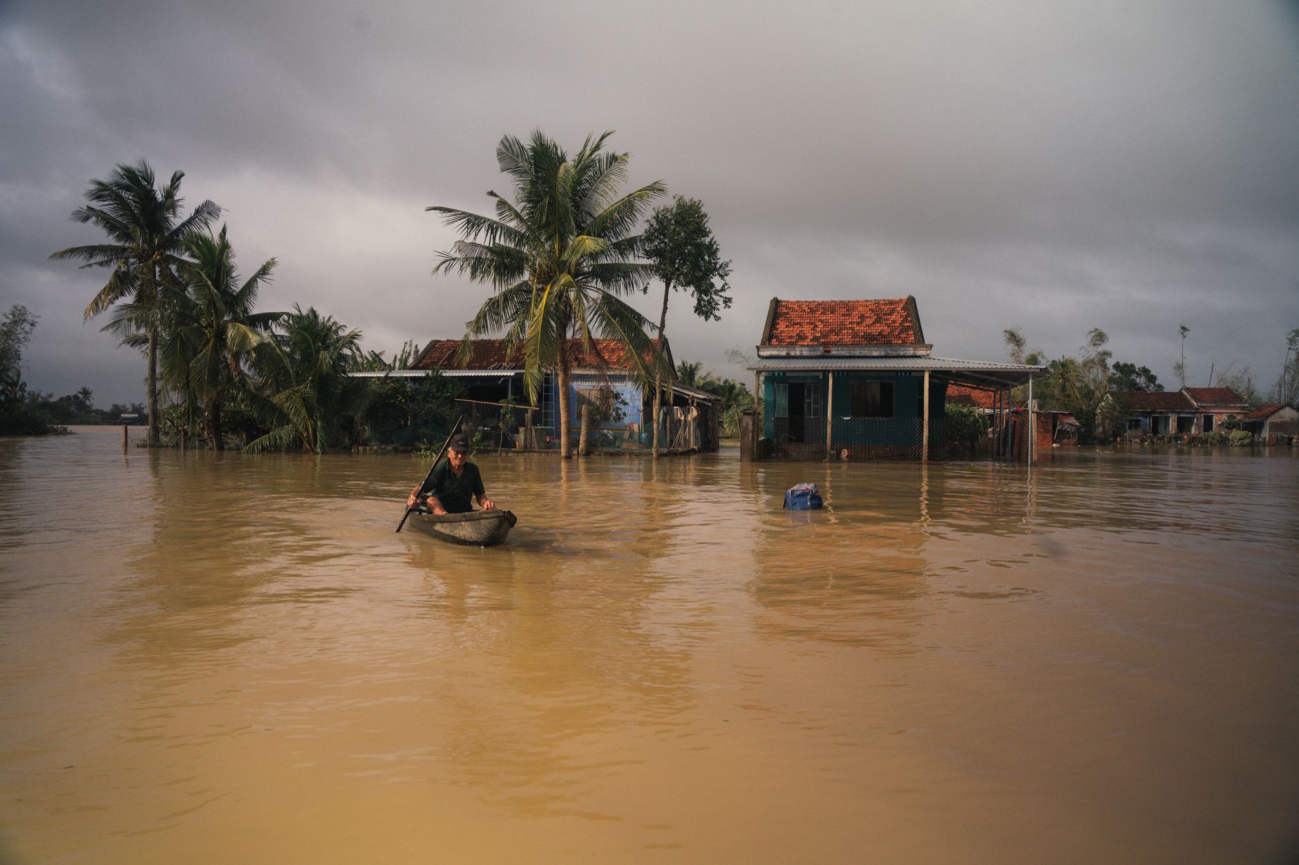 A man paddles a boat through a flooded village in Phú Yên, Vietnam, under cloudy skies.