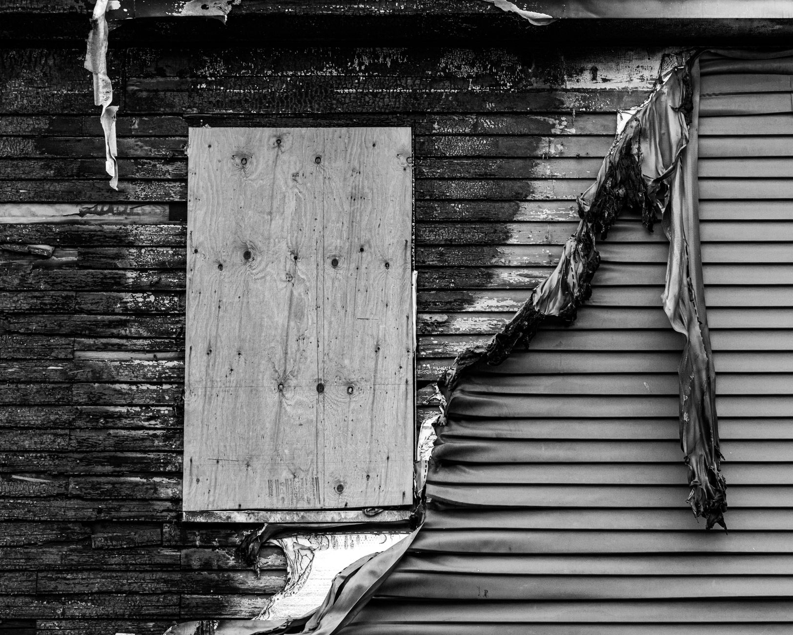 Artistic black and white photo displaying damaged exterior of a building, with charred wood and peeling siding.