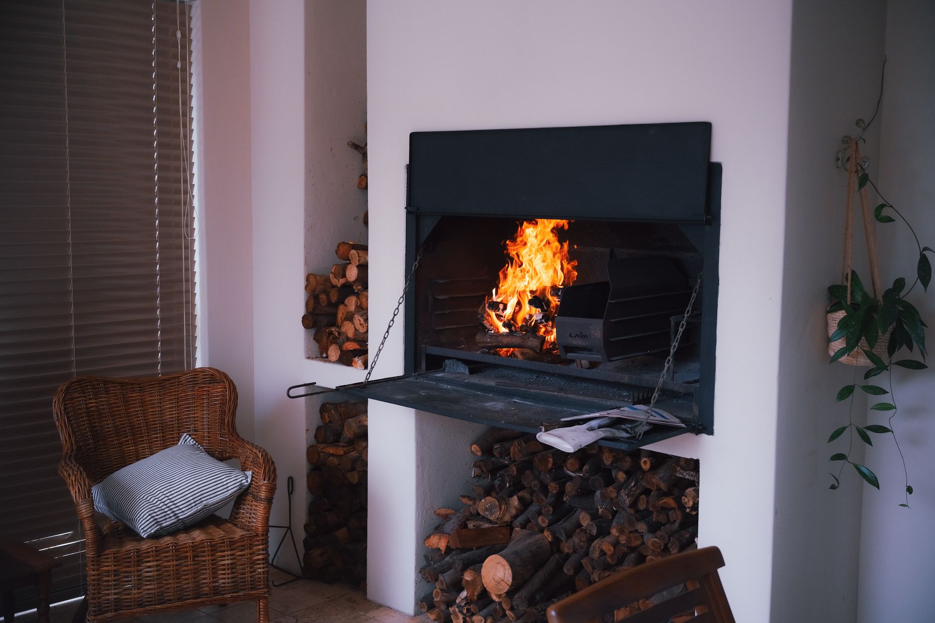 A warm and inviting indoor fireplace with stacked firewood, wicker chair, and plants for a cozy ambiance.