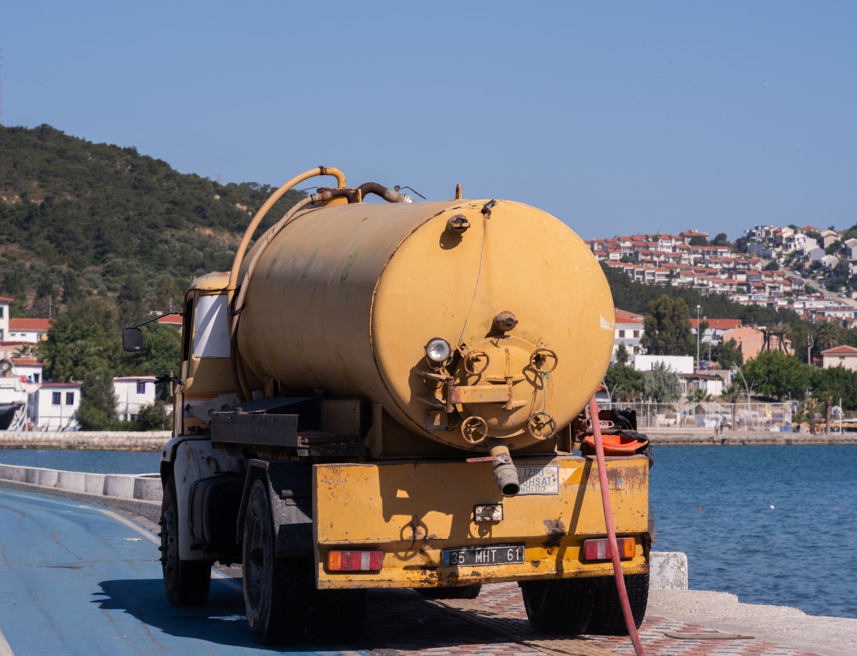 A yellow tanker truck parked by the harbor in Foça, İzmir, with a scenic coastal view.